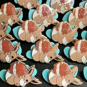 May include: A collection of enamel pins featuring a cartoon character with brown, white, and copper tones. The pins are set on light blue heart-shaped backings. The pins have a shiny, metallic finish.