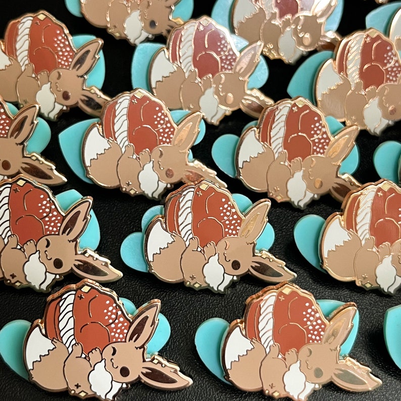 May include: A collection of enamel pins featuring a cartoon character with brown, white, and copper tones. The pins are set on light blue heart-shaped backings. The pins have a shiny, metallic finish.
