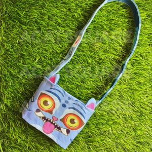 Könnte beinhalten: Eine hellblaue Umhängetasche mit einem Cartoon-Katzen-Gesicht-Design. Die Katze hat große gelbe Augen, eine rosa Zunge und rosa Ohren. Die Tasche hat einen passenden blauen Riemen und befindet sich vor einem grünen Gras-Hintergrund.