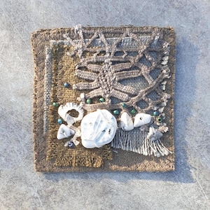 May include: A small square textile art piece with a brown background. The piece is made of different fabrics and textures, including burlap, lace, and yarn. It is decorated with white shells and small green beads.