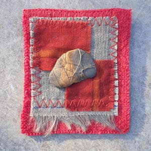 May include: A small patchwork quilt with red and grey fabric squares. A brown stone is placed on top of the quilt.