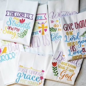 May include: A set of six white kitchen towels with colorful embroidered religious quotes. The quotes include "The Lord is my strength and my song", "Give thanks to the Lord for he is good", "Just enough grace for today", "Be still and know that I am God", "Whatever you do, do it for the glory of God", and "He who sings, prays twice".
