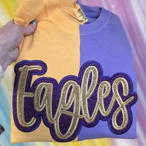 Könnte beinhalten: Ein zweifarbiges Sweatshirt, vertikal geteilt mit einem gelb-orangen und lila Farbschema. Das Wort "Eagles" ist in goldfarbenem Glitzer mit einem lila Umriss gestickt. Das Sweatshirt wird von einer Hand gehalten.