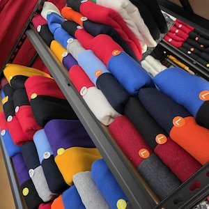 May include: A display of folded sweatshirts in various colors, including red, blue, black, and gray, arranged on shelves. Each sweatshirt has a size label, such as M, L, or XXL. The sweatshirts are neatly stacked, showcasing a variety of options.