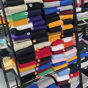 May include: A multi-tiered metal rack displays stacks of folded sweatshirts in various colors, including black, red, blue, and yellow. Each sweatshirt is folded neatly, with size labels visible. The sweatshirts are organized by color and size.