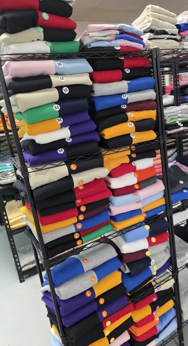 May include: A multi-tiered metal rack displays stacks of folded sweatshirts in various colors, including black, red, blue, and yellow. Each sweatshirt is folded neatly, with size labels visible. The sweatshirts are organized by color and size.