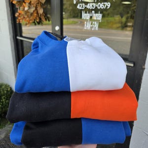 May include: A stack of four folded sweatshirts in contrasting colors: blue, white, black, and orange. Each sweatshirt is folded in half, with the colors split vertically. The sweatshirts are stacked on top of each other.