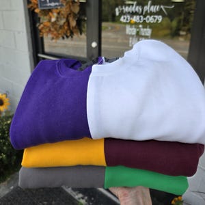 May include: A stack of folded crewneck sweatshirts in various colors: purple, white, gold, burgundy, gray, and green. The sweatshirts are neatly arranged, showcasing their solid colors. The image is taken outdoors, with a storefront visible in the background.