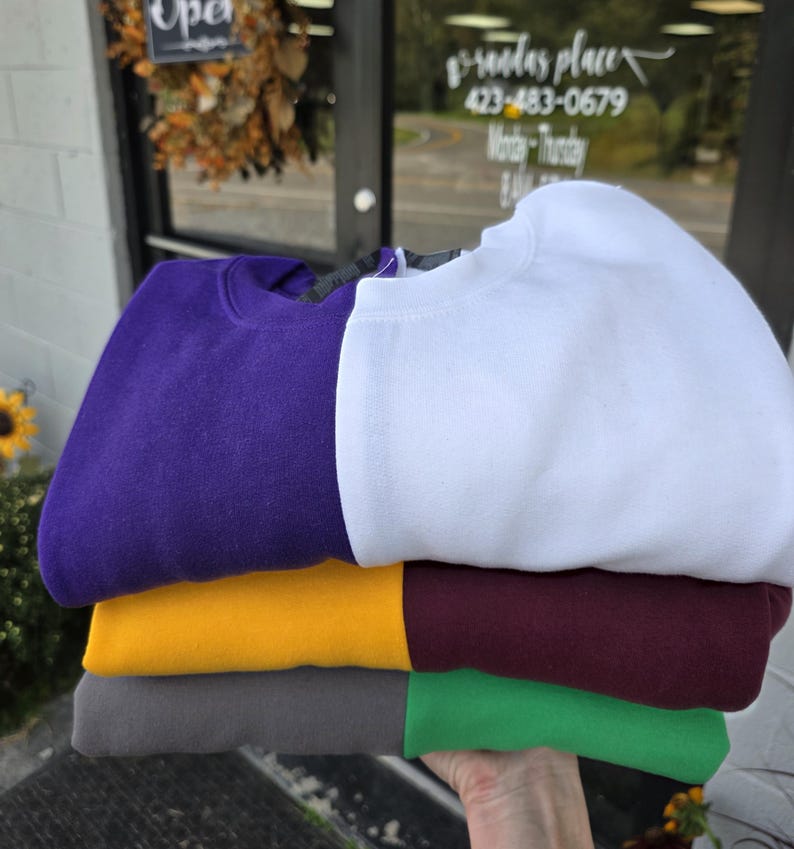 May include: A stack of folded crewneck sweatshirts in various colors: purple, white, gold, burgundy, gray, and green. The sweatshirts are neatly arranged, showcasing their solid colors. The image is taken outdoors, with a storefront visible in the background.
