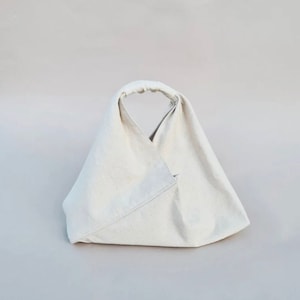 May include: A white canvas tote bag with a simple design. The bag has a single handle and is folded into a triangular shape.