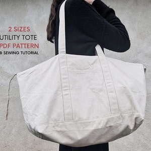 May include: A large, off-white canvas tote bag with two long straps. The bag has a single pocket on the front. The text "2 SIZES UTILITY TOTE PDF PATTERN & SEWING TUTORIAL" is printed in red on a white background.