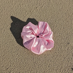 Puede incluir: Un scrunchie de satén rosa, una liga para el cabello, sobre una superficie arenosa.