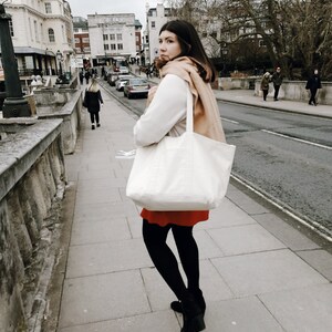 Peut inclure: Une femme portant un pull blanc, une jupe rouge et des collants noirs marche sur un trottoir en portant un grand sac fourre-tout blanc avec une &eacute;charpe brune drap&eacute; sur son &eacute;paule.