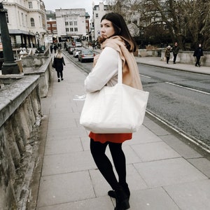 Puede incluir: Una mujer con un su&eacute;ter blanco, una falda roja y medias negras camina por una calle de la ciudad. Lleva un bolso tote blanco grande con una bufanda marr&oacute;n drapeada en la parte superior. La calle est&aacute; bordeada de edificios y &aacute;rboles.