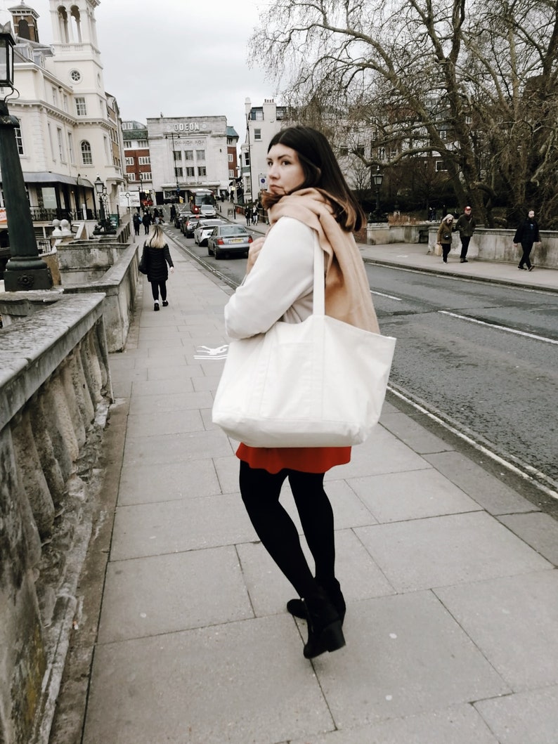 Puede incluir: Una mujer con un su&eacute;ter blanco, una falda roja y medias negras camina por una calle de la ciudad. Lleva un bolso tote blanco grande con una bufanda marr&oacute;n drapeada en la parte superior. La calle est&aacute; bordeada de edificios y &aacute;rboles.