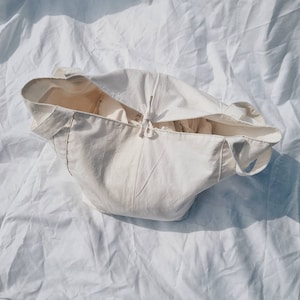 Peut inclure: Un sac fourre-tout en toile blanche avec une fermeture &agrave; cordon. Le sac est pli&eacute; et pos&eacute; sur une surface blanche.