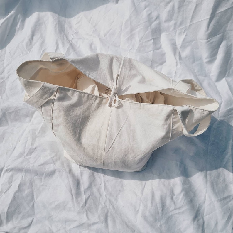 Peut inclure: Un sac fourre-tout en toile blanche avec une fermeture &agrave; cordon. Le sac est pli&eacute; et pos&eacute; sur une surface blanche.