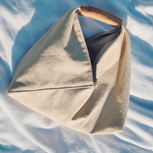 May include: A beige canvas tote bag with a brown leather handle. The bag is folded and has a triangular shape.