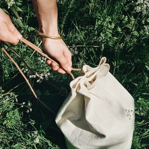 Könnte beinhalten: Eine weiße Canvas-Kordeltasche mit einem braunen Lederkordelzug. Die Tasche liegt auf einem Bett aus grünem Gras und weißen Blumen.