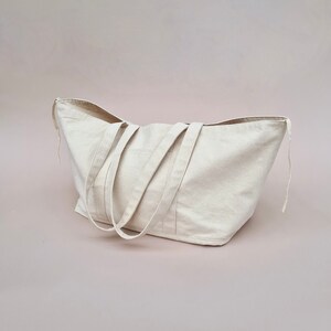 Puede incluir: Una bolsa de lona grande de color blanco roto con dos asas largas. La bolsa est&aacute; sobre una superficie rosa claro.