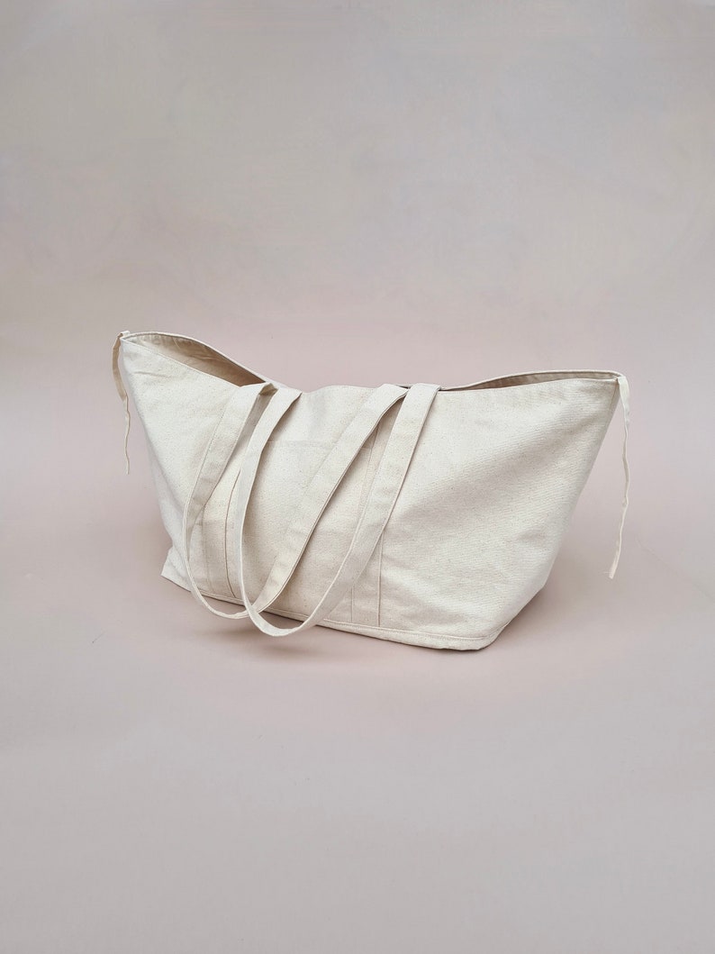 Puede incluir: Una bolsa de lona grande de color blanco roto con dos asas largas. La bolsa est&aacute; sobre una superficie rosa claro.