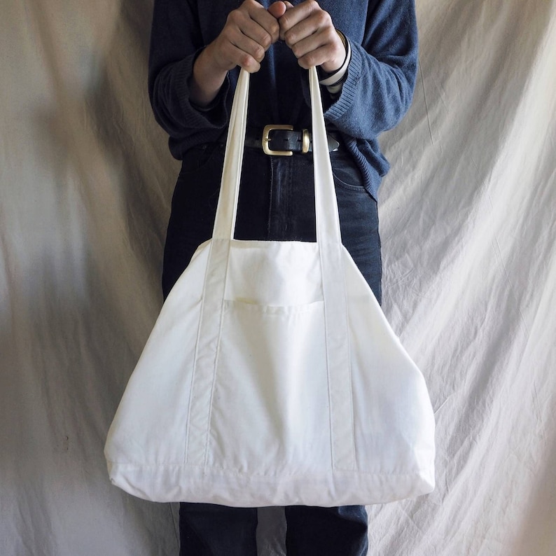 Puede incluir: Una bolsa de lona blanca con un bolsillo &uacute;nico en la parte delantera. La bolsa tiene dos correas largas y la sostiene una persona.