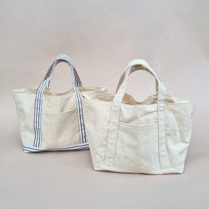 Peut inclure: Deux sacs cabas en toile beige avec des anses ray&eacute;es. Un sac a une seule poche sur le devant, tandis que l'autre a deux poches.