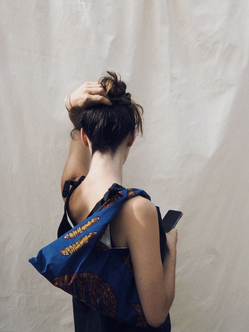 May include: A woman with her hair pulled back in a bun is wearing a blue and yellow patterned tote bag over her shoulder. She is holding a black smartphone in her right hand.