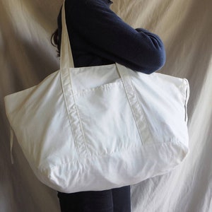Puede incluir: Una gran bolsa de lona blanca con un solo bolsillo en la parte delantera. La bolsa tiene asas largas y un cierre de cord&oacute;n.