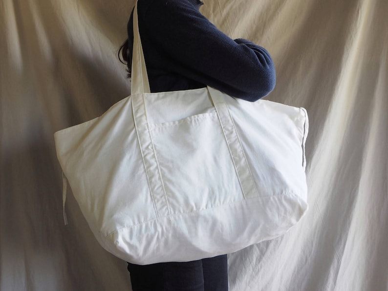 Puede incluir: Una gran bolsa de lona blanca con un solo bolsillo en la parte delantera. La bolsa tiene asas largas y un cierre de cord&oacute;n.