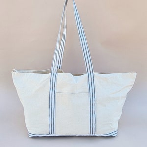 Puede incluir: Una bolsa de lona blanca con asas rayadas azules y negras. La bolsa tiene un compartimento principal grande y un bolsillo m&aacute;s peque&ntilde;o en la parte delantera.