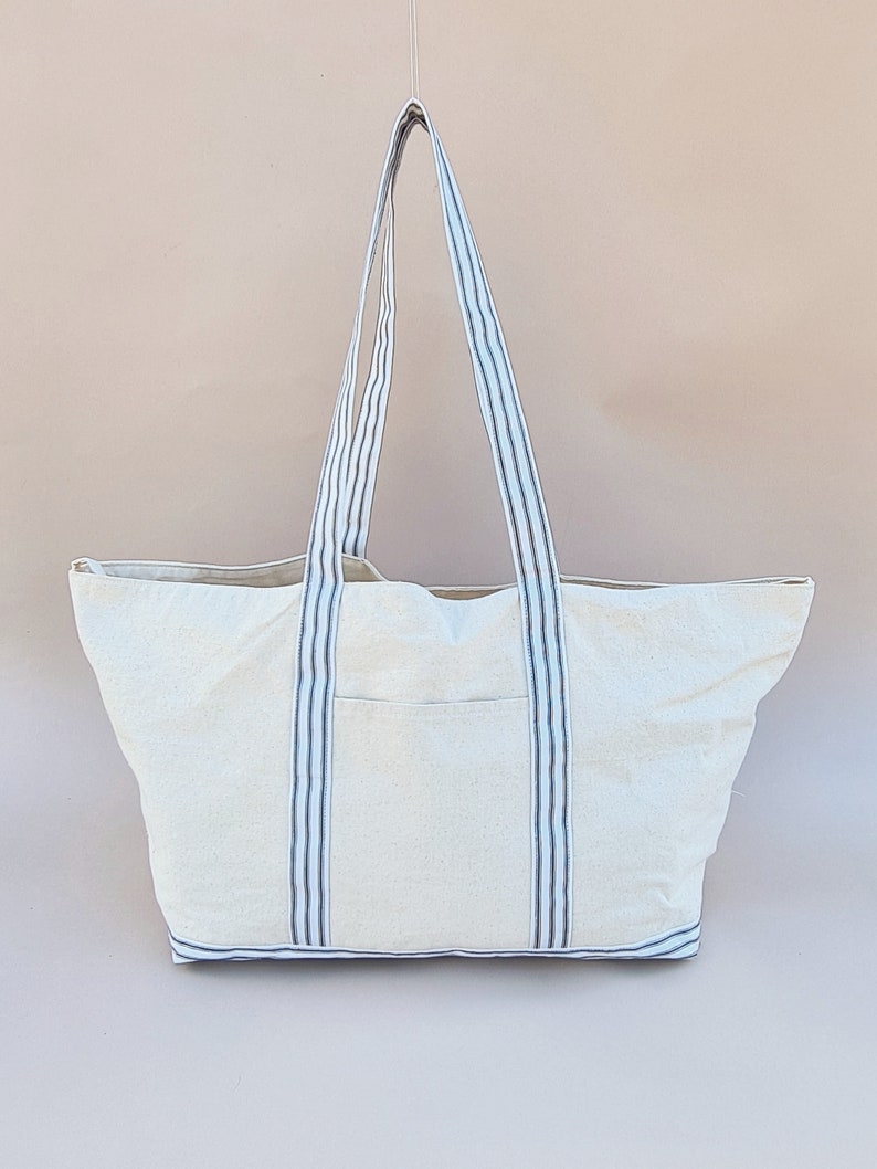 Puede incluir: Una bolsa de lona blanca con asas rayadas azules y negras. La bolsa tiene un compartimento principal grande y un bolsillo m&aacute;s peque&ntilde;o en la parte delantera.