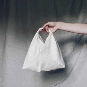May include: A white canvas tote bag with a triangular shape and a single handle. The bag is held by a hand in the image.