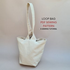 May include: A cream-colored loop bag with a single handle, suspended against a neutral background. The bag's design is simple, with a wide opening and a sturdy handle. Text on the right reads: "LOOP BAG PDF SEWING PATTERN & SEWING TUTORIAL."