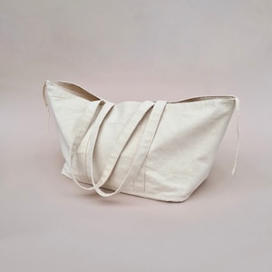 Peut inclure: Un grand sac fourre-tout en toile de couleur cr&egrave;me avec deux longues anses. Le sac est pos&eacute; sur une surface rose p&acirc;le.