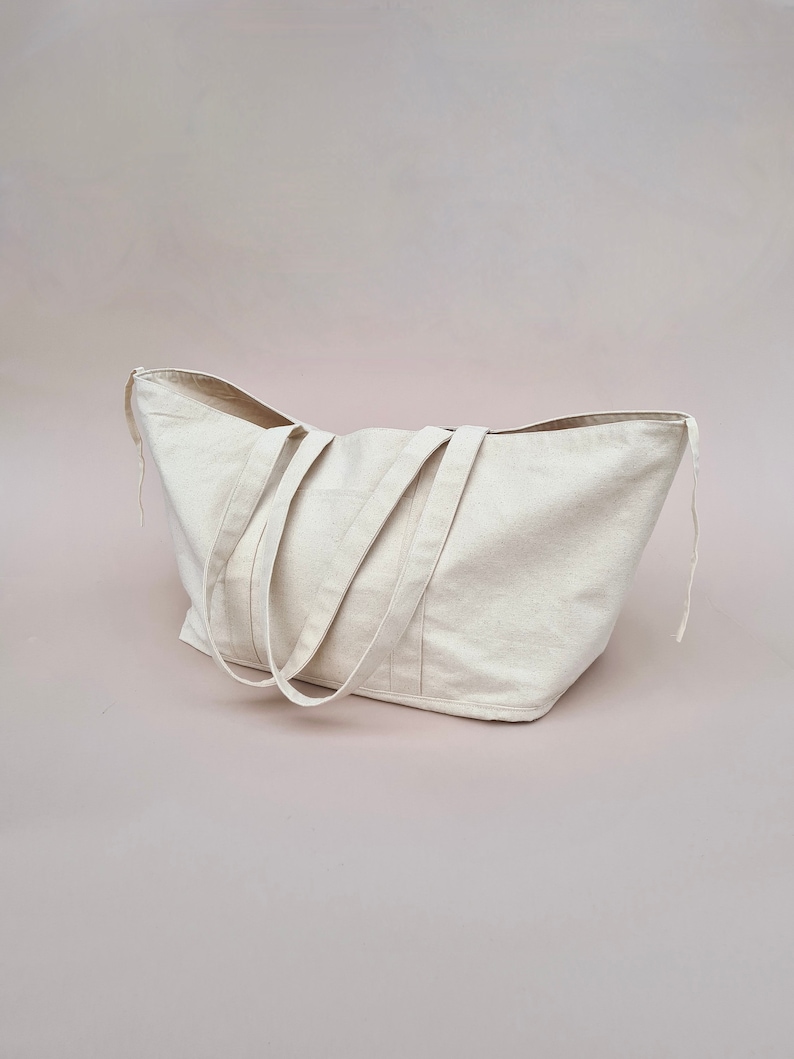Peut inclure: Un grand sac fourre-tout en toile de couleur cr&egrave;me avec deux longues anses. Le sac est pos&eacute; sur une surface rose p&acirc;le.