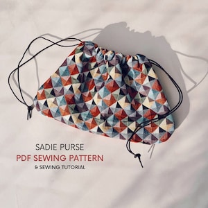 Pode incluir: Uma bolsa de cordão com um padrão geométrico em tons de vermelho, azul, verde e amarelo. A bolsa é feita de tecido e tem um cordão de ajuste preto. O texto "SADIE PURSE PDF SEWING PATTERN & SEWING TUTORIAL" está impresso abaixo da bolsa.