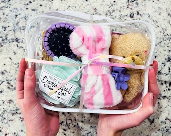May include: A clear, rectangular cosmetic bag filled with self-care items. Includes a purple hairbrush, a pink and white striped robe, a small teddy bear, a loofah, and a tag that reads "You are Beautiful inside & out!"