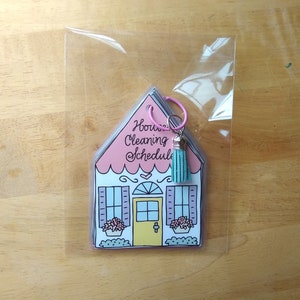 Op de afbeelding: Een roze en witte huisvormige schoonmaakschema met een turquoise kwast bevestigd aan een zilveren ring. De tekst op het huis luidt "House Cleaning Schedule".