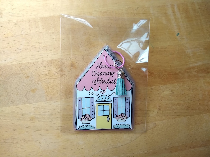 Op de afbeelding: Een roze en witte huisvormige schoonmaakschema met een turquoise kwast bevestigd aan een zilveren ring. De tekst op het huis luidt "House Cleaning Schedule".