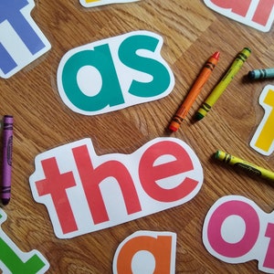 Può includere: Un set di flashcard di parole visive colorate e rivestite in schiuma con parole come "it", "as", "the", "of" e "to". Le flashcard sono su una superficie di legno con pastelli.