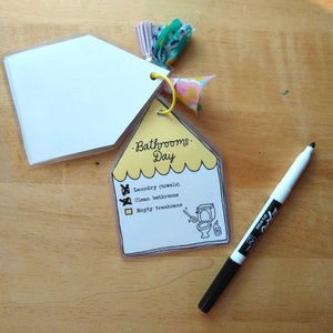Op de afbeelding: Een gele checklist met de tekst "Bathrooms Day" en een lijst met taken: "Laundry (towels)", "Clean bathrooms" en "Empty trashcans". De checklist is bevestigd aan een witte kaart met een decoratieve kwast.