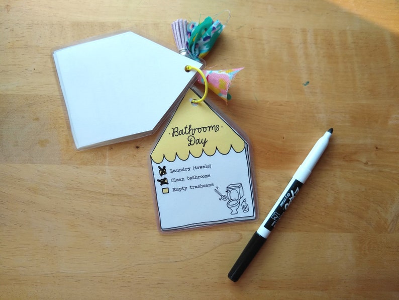 Op de afbeelding: Een gele checklist met de tekst "Bathrooms Day" en een lijst met taken: "Laundry (towels)", "Clean bathrooms" en "Empty trashcans". De checklist is bevestigd aan een witte kaart met een decoratieve kwast.