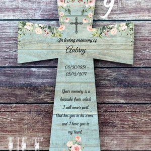Puede incluir: Una cruz de madera con fondo azul claro y detalles florales. La cruz tiene una cinta blanca colgando de la parte superior. El texto en la cruz dice: "In loving memory of Aubrey 08/30/1951-05/12/2017 Your memory is a keepsake from which I will never part. God has you in his arms, and I have you in my heart."