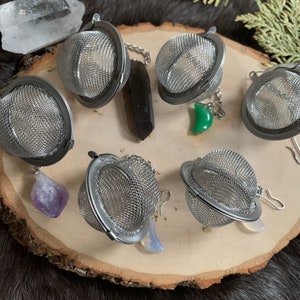 May include: Six silver tea infusers with mesh balls and chain handles. The infusers are arranged on a wooden surface with a few crystals and stones.