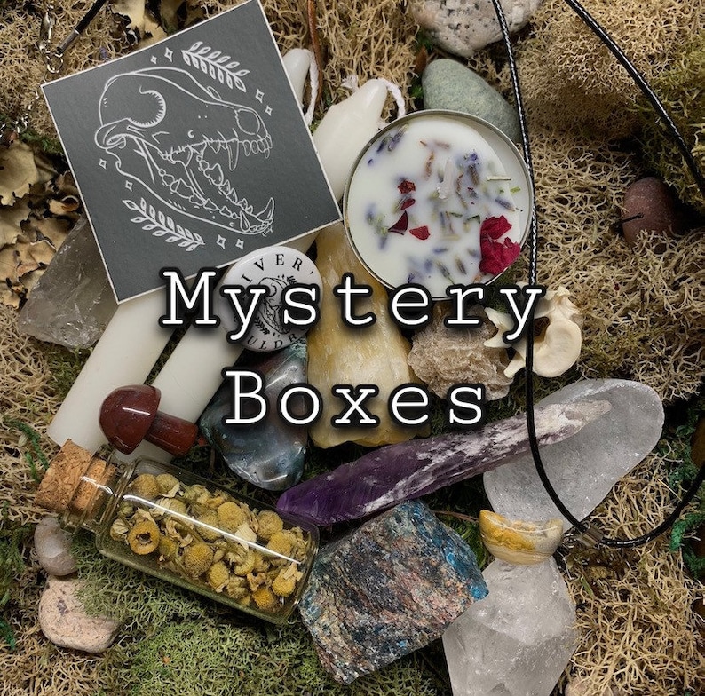 May include: A collection of items including crystals, candles, and a small bottle filled with dried flowers, arranged on a bed of moss. A card with a skull illustration and the words "Mystery Boxes" are visible. The items are in various colors and textures.