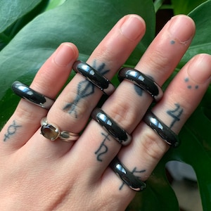 May include: A hand wearing six silver rings, each with a smooth, polished finish. The rings are stacked on each finger, creating a bold and modern look.