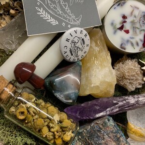 May include: An assortment of crystals, candles, and botanicals. A small glass bottle filled with dried chamomile flowers sits next to a red mushroom-shaped stone. A button with the text "River's Cauldron" is also visible.
