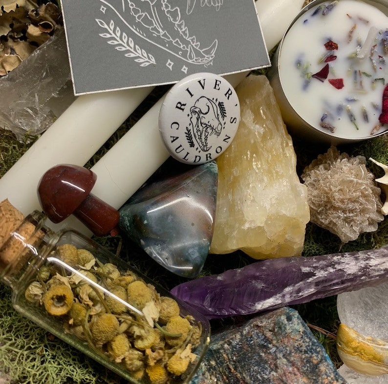 May include: An assortment of crystals, candles, and botanicals. A small glass bottle filled with dried chamomile flowers sits next to a red mushroom-shaped stone. A button with the text "River's Cauldron" is also visible.