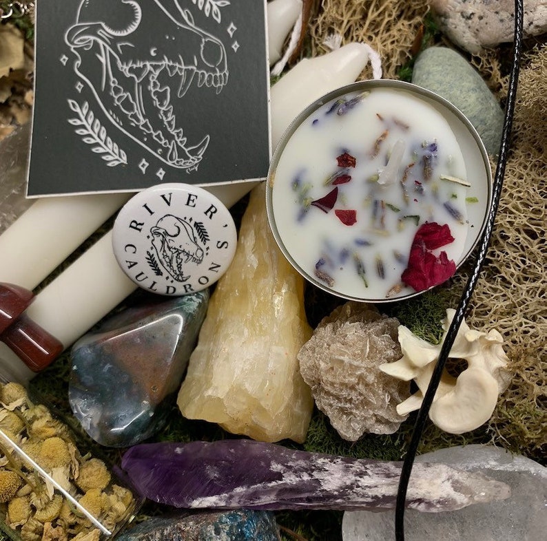 May include: An assortment of crystals, a small candle with dried flowers, and a button that says "River's Cauldron" with a skull graphic. The items are arranged on a bed of moss and dried flowers, with a black cord visible.
